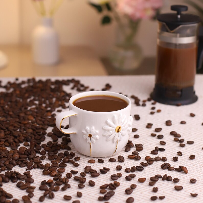 May include: A white ceramic mug filled with dark coffee, featuring embossed white daisy designs with gold centers and gold-trimmed handles, surrounded by scattered roasted coffee beans and a black French press.