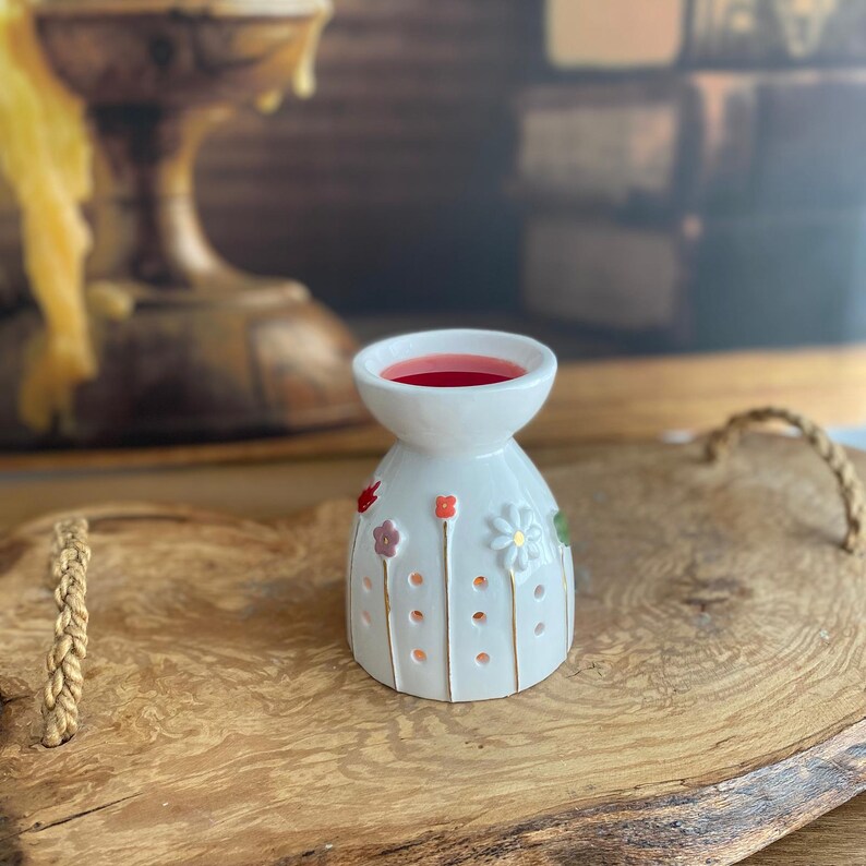 May include: A white ceramic wax warmer with a bell-shaped body featuring small, raised, colorful flower motifs and thin gold vertical lines. The top bowl contains a pool of deep red wax, and the base has small circular cutouts for light.