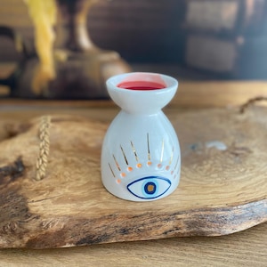 May include: A white ceramic oil burner with a flared top and a rounded base. The exterior features a painted blue and gold eye motif with radiating gold lines above it. The interior of the top basin contains a small amount of red wax.