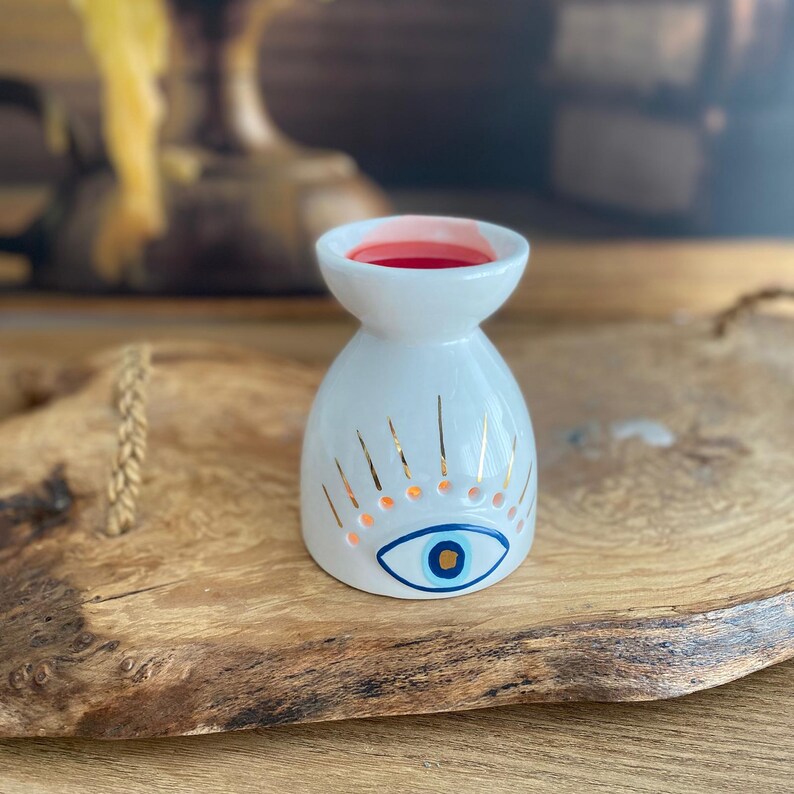 May include: A white ceramic oil burner with a flared top and a rounded base. The exterior features a painted blue and gold eye motif with radiating gold lines above it. The interior of the top basin contains a small amount of red wax.