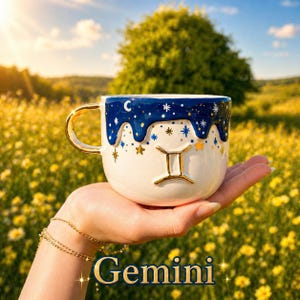 May include: A white ceramic mug featuring a gold Gemini zodiac symbol and a dark blue drip-style rim decorated with gold and white stars and a crescent moon. The mug has a gold-colored handle and a smooth, glossy finish on the exterior surface.