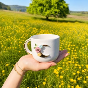 May include: White ceramic mug with a gold handle and a crescent moon design adorned with colorful flowers. The mug is held in a hand, set against a backdrop of a field of yellow flowers and a tree.
