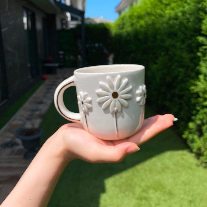 May include: A white ceramic mug featuring embossed daisy flower designs on the exterior. The mug has a curved handle with a thin gold-colored trim along the edge. The surface of the mug is smooth and glossy, highlighting the raised floral petals.
