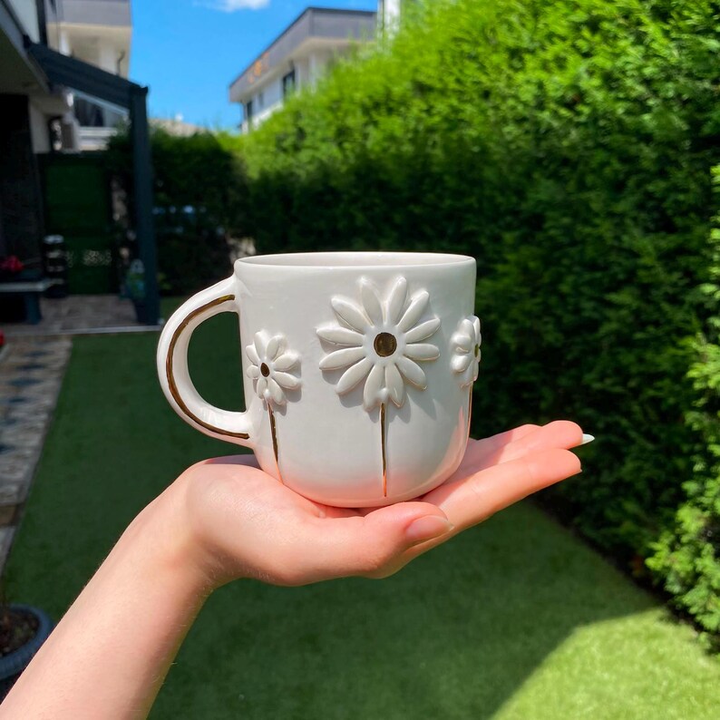 May include: A white ceramic mug featuring raised, three-dimensional daisy flower designs with gold-painted centers and stems. The mug has a curved handle with a gold-painted edge, providing a decorative and tactile finish to the exterior surface.