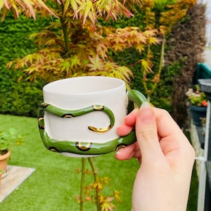 May include: A white ceramic mug featuring a sculpted green snake wrapped around the exterior. The snake has a smooth, glossy finish with gold-painted accents along its body, forming the handle and decorative elements on the mug's surface.