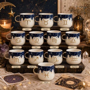 May include: A collection of twelve ceramic mugs featuring a dark blue starry night sky design dripping over a cream-colored base. Each mug displays a unique gold-colored zodiac symbol, with corresponding labels for Aries, Taurus, Gemini, Cancer, Leo, Virgo, Libra, Scorpio, Sagittarius, Capricorn, Aquarius, and Pisces.
