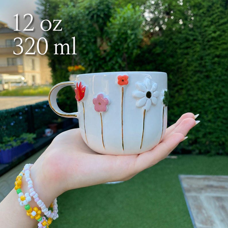 May include: White ceramic mug with a gold handle and floral embellishments. The mug features raised, colorful flowers with gold stems. The text "12 oz" and "320 ml" is printed on the mug.