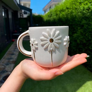 May include: A white ceramic mug featuring embossed daisy flower designs on the exterior. The mug has a curved handle with a gold-colored trim, and the centers of the daisy petals are accented with small, reflective gold-colored circular details.