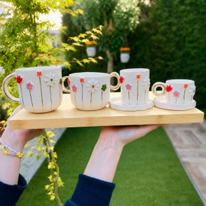 May include: A set of four white ceramic mugs and cups with floral designs, presented on a bamboo tray. Each piece features gold accents on the handles and stems, with colorful, raised flower details. The set includes a large mug, two smaller mugs, and a cup with a saucer.