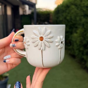 May include: A white ceramic mug featuring three raised, sculpted daisy flowers with white petals and brown centers. The mug has a gold-trimmed handle and thin gold lines extending downward from each flower to the base of the mug.