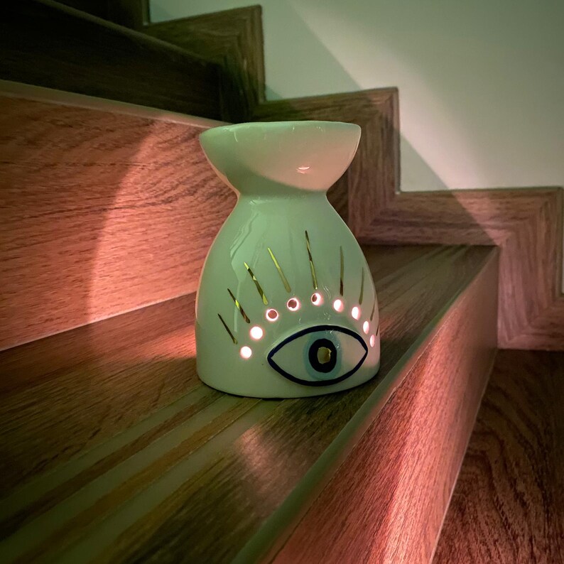 May include: A white ceramic oil burner featuring a stylized eye design with gold-colored eyelashes and a central blue iris. The body of the burner has small circular cutouts that allow light to shine through from the interior, creating a glow.