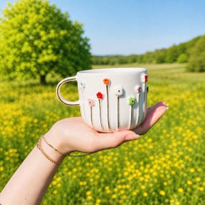May include: A white ceramic mug with a gold handle and rim, adorned with colorful, raised flower designs. The mug is held in a hand against a backdrop of a green field and blue sky. The flowers are red, orange, pink, and green.