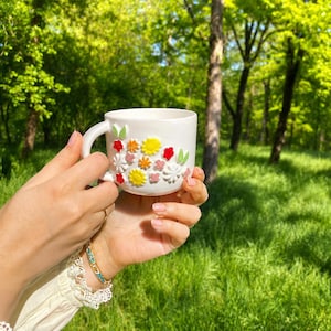 May include: White ceramic mug with a handle, adorned with colorful, raised floral designs. The flowers include red, yellow, orange, and white blossoms with green leaves. The mug is held against a backdrop of green foliage.
