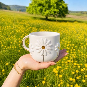 May include: White ceramic mug with embossed daisy flower designs and gold accents. The mug is held in a hand, set against a backdrop of a field of yellow flowers and a tree.