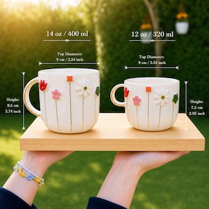 May include: Two white ceramic mugs with floral designs. The larger mug is 3.74 inches tall and holds 14 oz. The smaller mug is 2.95 inches tall and holds 12 oz. Both mugs have a top diameter of 3.54 inches.