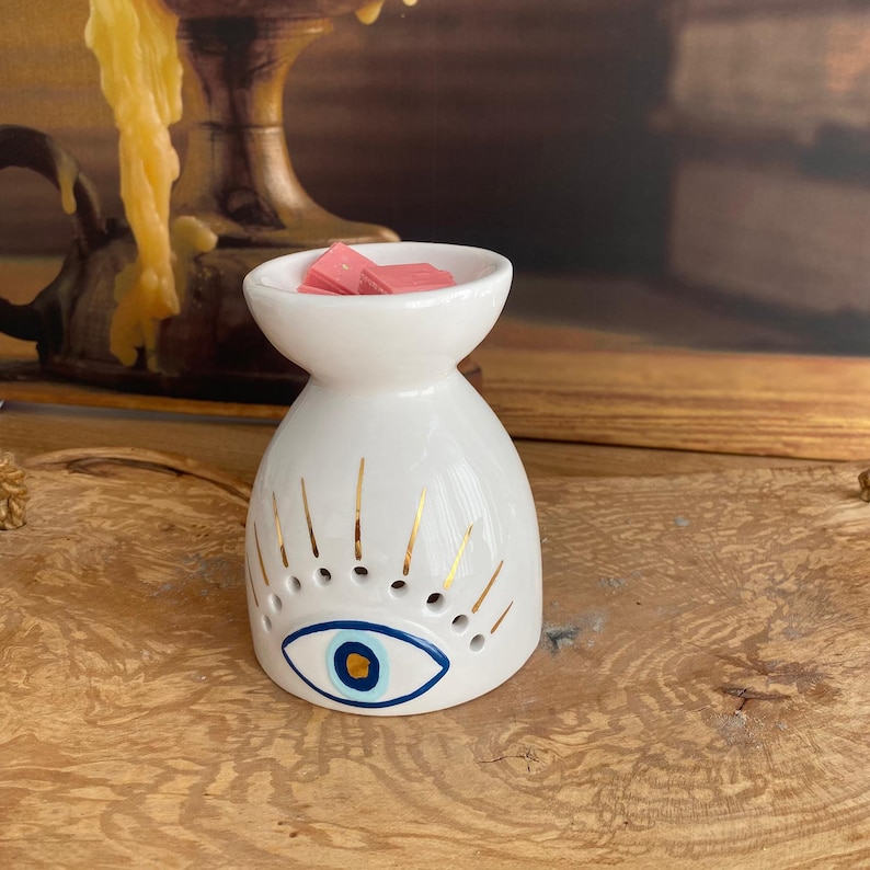 May include: A white ceramic wax warmer featuring a painted blue and gold eye design on the front. The top bowl contains small pink wax melts. The body of the warmer has a rounded shape with gold painted eyelashes radiating from the central eye motif.