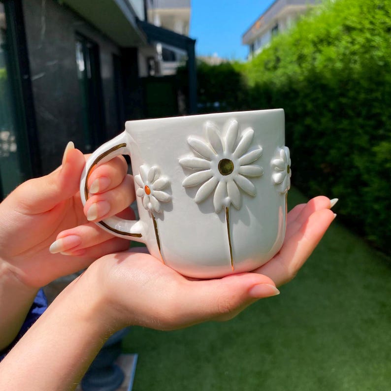 May include: A white ceramic mug featuring raised, three-dimensional daisy flower designs on the exterior. The petals are white with gold-painted stems and centers, and the handle is accented with a thin gold trim along the outer edge.