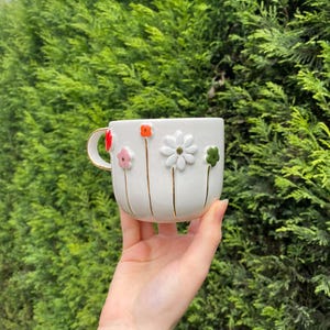 May include: White ceramic mug with a gold handle and rim, adorned with colorful 3D flower designs. The flowers include red, pink, orange, white, and green, with gold stems. The mug is held against a backdrop of green foliage.
