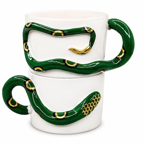 May include: Two white ceramic mugs stacked vertically, each featuring a three-dimensional green snake wrapped around the exterior. The snakes have gold-painted accents along their bodies and textured scales on their heads, with the tail forming the handle.