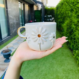 May include: A white ceramic mug featuring three-dimensional embossed daisy flowers on the exterior. The center of each daisy is a reflective gold-toned circle. The mug has a curved handle with a thin gold-colored rim along the top edge.