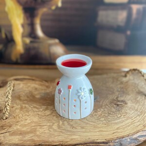 May include: A white ceramic wax warmer featuring a bell-shaped body decorated with small, colorful painted flowers on thin stems. The top bowl contains a pool of vibrant red wax, and the base is perforated with small circular holes for light.
