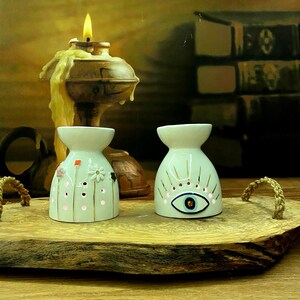 May include: Two white ceramic oil burners with flared tops. The left burner features small painted flowers and perforated dots. The right burner displays a central eye motif with radiating gold lines. Both items are illuminated from within.