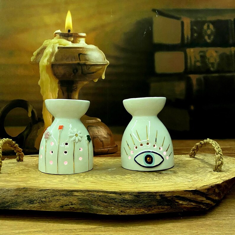 May include: Two white ceramic oil burners with flared tops. The left burner features small painted flowers and perforated dots. The right burner displays a central eye motif with radiating gold lines. Both items are illuminated from within.