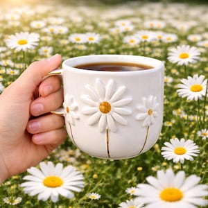 May include: White ceramic mug with embossed daisy flowers and gold accents, filled with a dark beverage. The mug is held in a hand, set against a backdrop of a field of daisies.