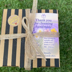 May include: A square cardboard gift box with black and tan vertical stripes, tied with a natural jute twine bow. A circular sticker reads Hand Made With Love. A rectangular card features floral illustrations and text: Thank you for choosing handmade.