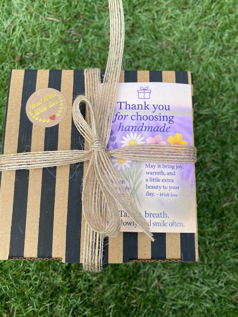 May include: A square cardboard gift box with black and tan vertical stripes, tied with a natural jute twine bow. A circular sticker reads Hand Made With Love. A rectangular card features floral illustrations and text: Thank you for choosing handmade.