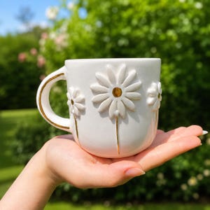 May include: A white ceramic mug featuring raised, three-dimensional daisy flower designs on the exterior. The mug includes gold-painted accents on the flower centers, the stems, and the edges of the handle, creating a decorative finish.