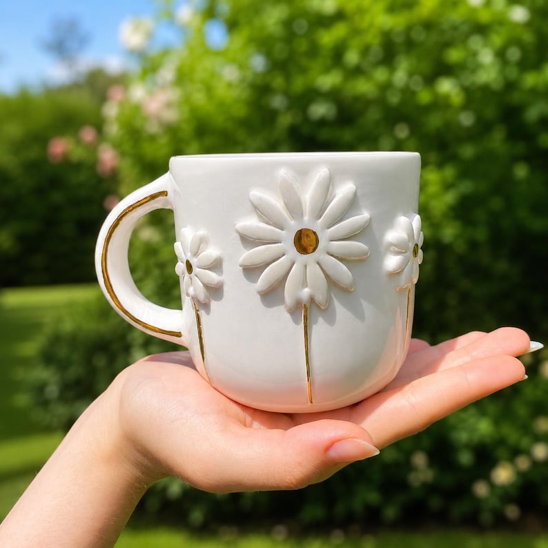 May include: A white ceramic mug featuring raised, three-dimensional daisy flower designs on the exterior. The mug includes gold-painted accents on the flower centers, the stems, and the edges of the handle, creating a decorative finish.