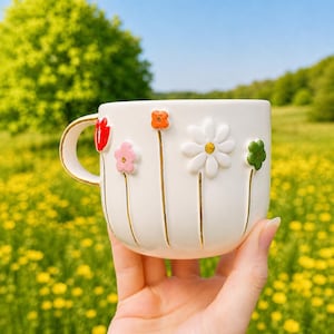 May include: A white ceramic mug with a gold handle and rim, adorned with embossed, colorful flowers. The flowers include a red tulip, pink flower, orange flower, white daisy, and green flower. The mug is held in front of a blurred green and yellow background.