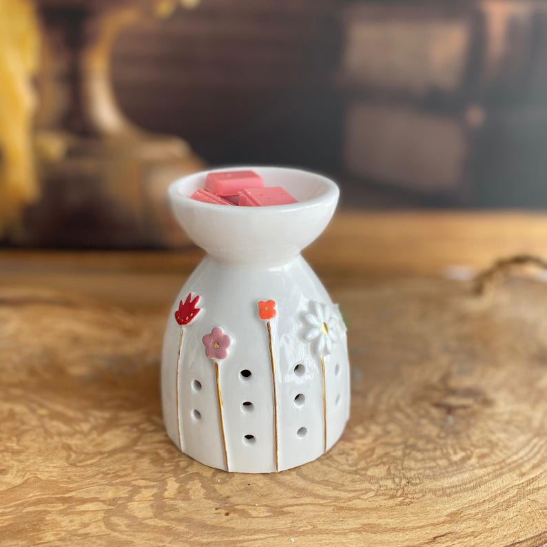 May include: A white ceramic wax warmer featuring a bowl top filled with pink wax melts. The exterior is decorated with painted flowers in red, pink, and orange, along with a white daisy relief. Vertical gold lines and small circular cutouts adorn the base.