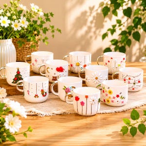 May include: A collection of white ceramic mugs with gold handles, each adorned with unique, colorful floral and seasonal designs. The mugs are arranged on a wooden surface, showcasing a variety of decorative patterns and textures.