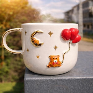 May include: A white ceramic mug with a gold handle and rim, decorated with a gold crescent moon, stars, and a teddy bear holding red balloons. The mug is filled with a hot beverage, and steam is rising from the top.