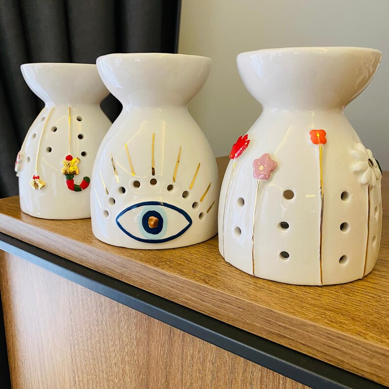 May include: Three white ceramic oil warmers with decorative designs. One features a gold and blue eye, another has Christmas-themed charms, and the third has floral accents. Each warmer has a wide top and a series of holes.