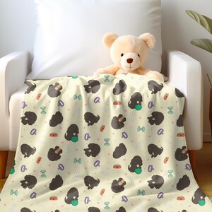 Può includere: Una coperta color crema con un motivo ripetuto di cani neri, palline verdi e illustrazioni di ossa e ciotole per cibo. La coperta è drappeggiata su una poltrona bianca, con un orsacchiotto sopra. Una pianta da interno in un cesto intrecciato è sullo sfondo.