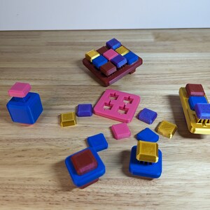 Könnte beinhalten: Ein Satz bunter 3D-gedruckter Puzzle-Teile in verschiedenen Formen und Größen. Die Teile sind aus blauem, rotem, goldenem und violettem Kunststoff. Die Teile sind auf einer Holzoberfläche angeordnet.
