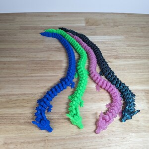 Puede incluir: Cinco juguetes de dragón de plástico flexibles, segmentados, en diferentes colores: azul, verde, rosa, negro y verde azulado. Los dragones están dispuestos en una línea curva, con sus cabezas mirando al espectador.