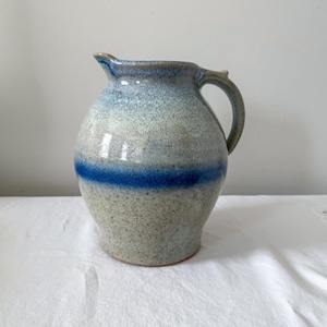 May include: A handcrafted ceramic jug with a rounded form and a handle. The jug has a speckled, light grey base with a vibrant blue stripe and a blue-toned rim. This jug is a decorative and functional item.