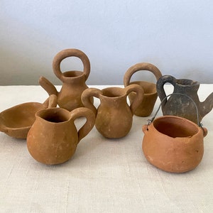 Puede incluir: Una colección de seis pequeñas piezas de cerámica de terracota, que incluyen una jarra, un tazón y una olla con asa. Las piezas son todas de varios tonos de marrón y tienen un aspecto rústico y hecho a mano.