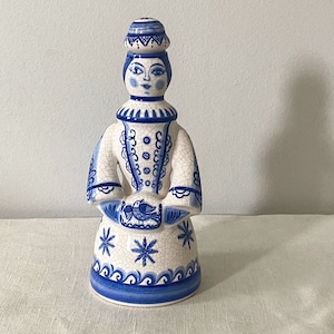 May include: A ceramic figurine of a woman wearing a blue and white patterned dress and hat. The figurine is hand-painted and has a detailed design.