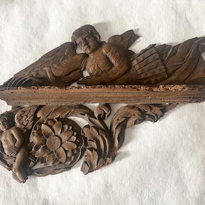 May include: A carved wooden shelf with two cherubs and a floral design. The shelf is made of dark wood and has a distressed finish.