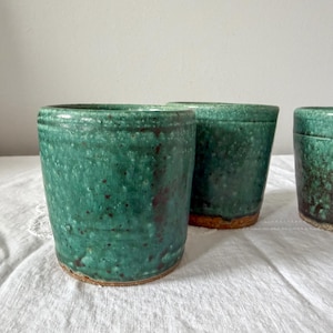 Puede incluir: Tres tazas de cerámica hechas a mano con un esmalte moteado de color verde azulado. Las tazas tienen forma cilíndrica con un diseño ligeramente cónico. La base de cada taza tiene un color arcilla natural sin esmaltar. Las tazas se exhiben sobre una superficie blanca.