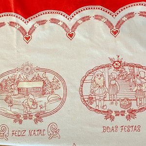 Puede incluir: Tela blanca con borde rojo y diseños bordados en rojo. Los diseños incluyen una escena navideña con Santa Claus y niños, una escena de invierno con una cabaña y el texto "Feliz Natal" y "Boas Festas".