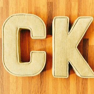 May include: Close-up of the letters "CK" in a light green corduroy fabric. The letters are outlined with a darker green stitching and sit on a wooden background. The letters are thick and stand out.