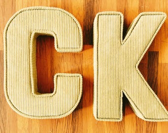 May include: Close-up of the letters "CK" in a light green corduroy fabric. The letters are outlined with a darker green stitching and sit on a wooden background. The letters are thick and stand out.
