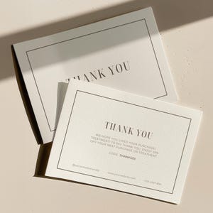 May include: Two off-white thank you cards with a simple black border and text. The top card reads "THANK YOU". The bottom card includes the text "THANK YOU", a message, and a discount code. The cards are on a light beige surface.