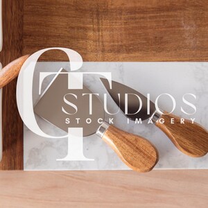 Puede incluir: Dos cuchillos de queso con mangos de madera y hojas plateadas sobre una superficie de mármol blanco. Los cuchillos están sobre una tabla de cortar de madera.